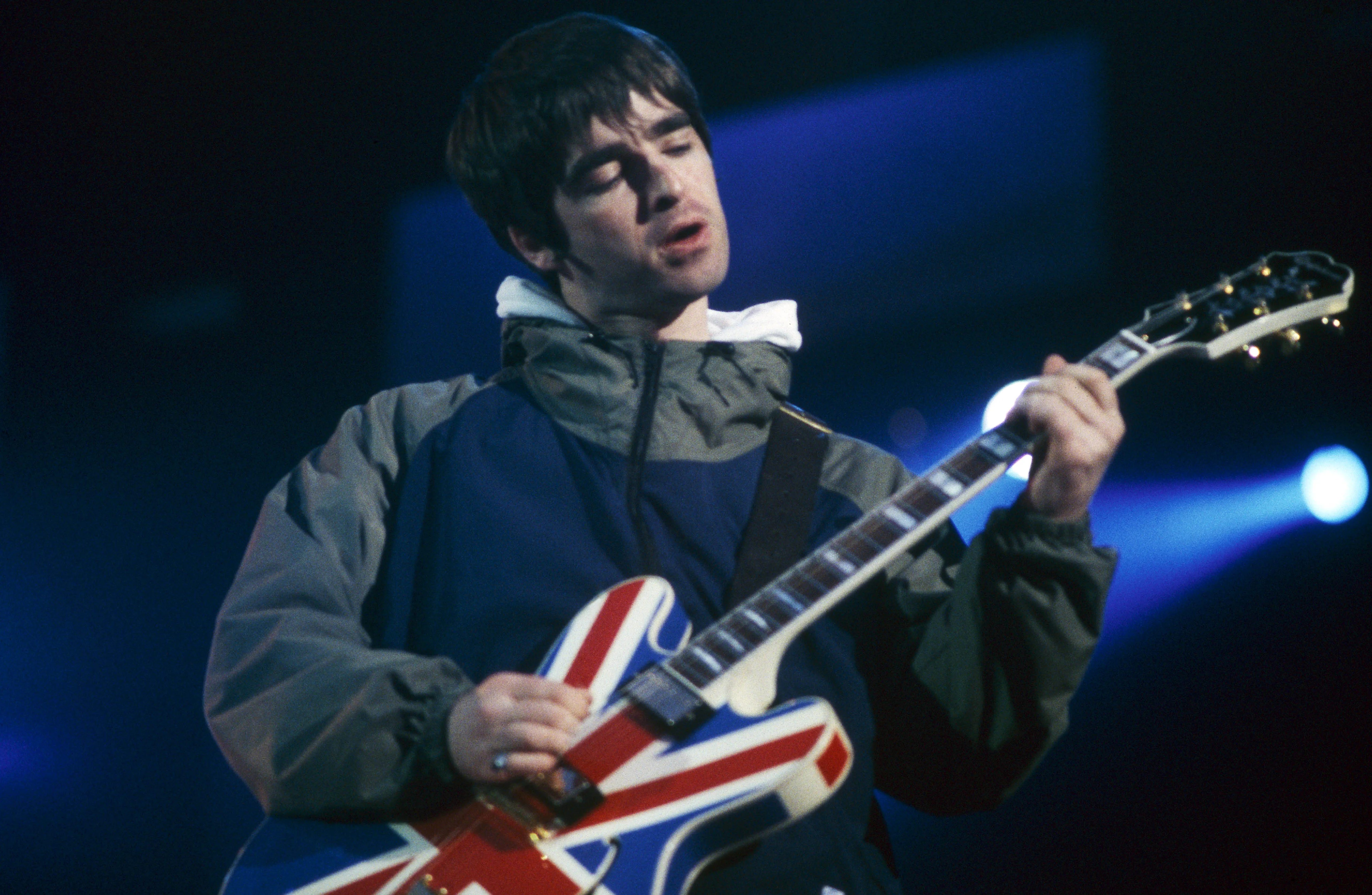 Noel Gallagher of Oasis wears a coat and plays a guitar with the Union Jack on it.