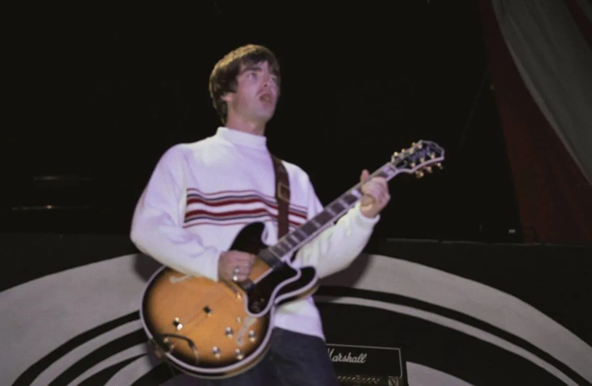 Noel Gallagher wears a white shirt and plays guitar.