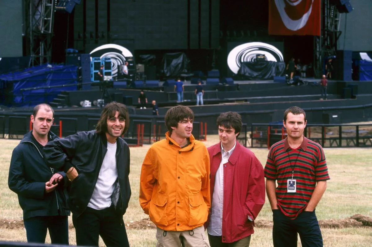  Paul 'Bonehead' Arthurs, Liam Gallagher, Noel Gallagher, Paul 'Guigsy' McGuigan and Alan White of Oasis stand in front of an outdoor stage.