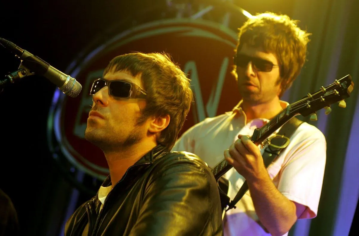 Liam and Noel Gallagher of Oasis perform on a stage together. They both wear sunglasses.