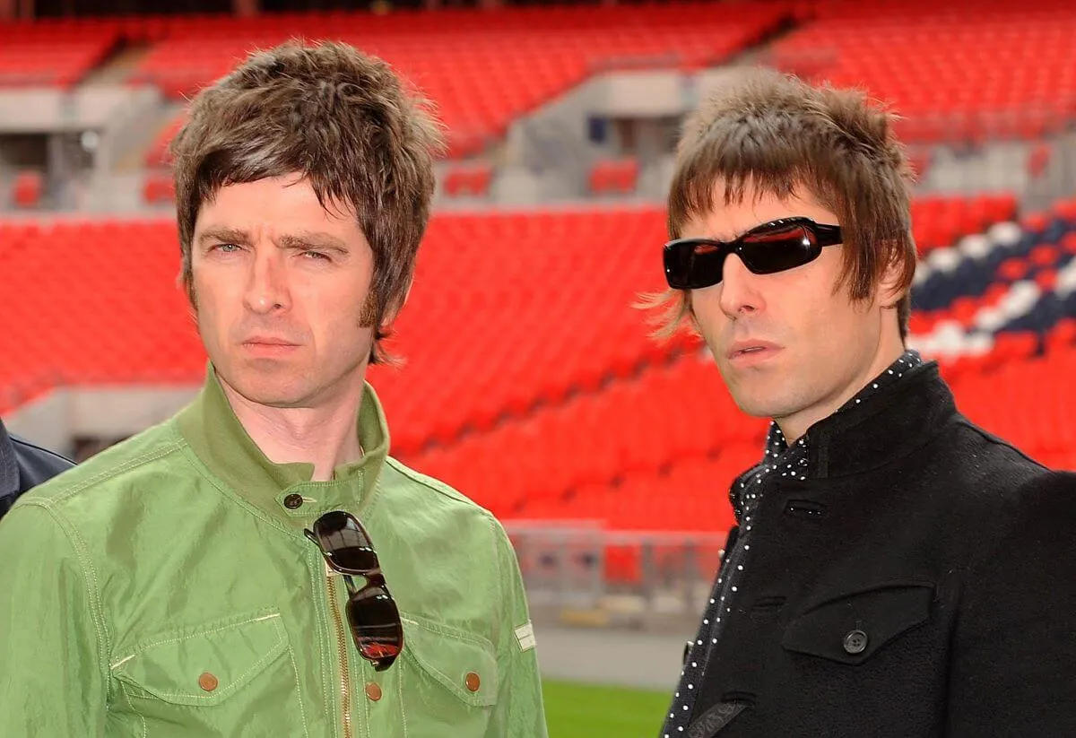 Noel Gallagher wears a green coat and stands next to Liam Gallagher, who wears a black coat. They stand in a football stadium.