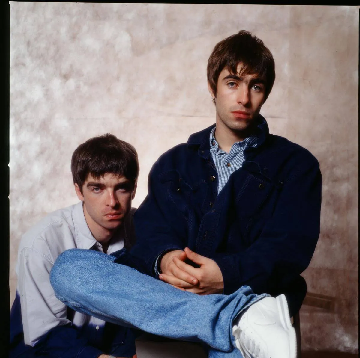 Noel and Liam Gallagher pose in front of a beige background. Noel crouches next to Liam, who sits in a chair with his legs up.