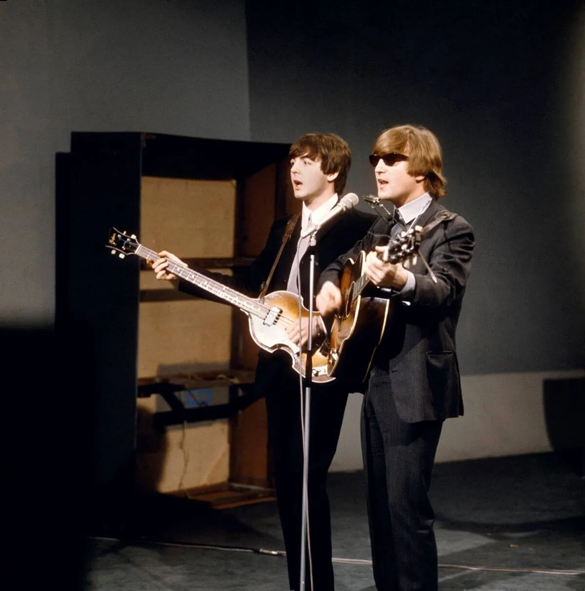 Paul McCartney and John Lennon wear suits and play guitar. Lennon wears sunglasses. They share a microphone.