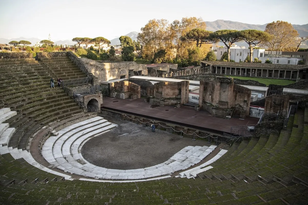 A broad view of Pompeii's Large Theater from above, with the ruins turning green from vegetation