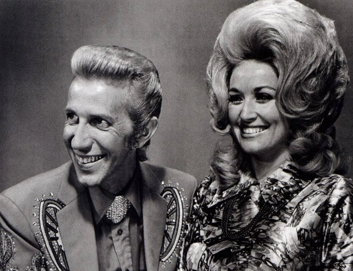 A black and white picture of Porter Wagoner and Dolly Parton posing together. They both smile.