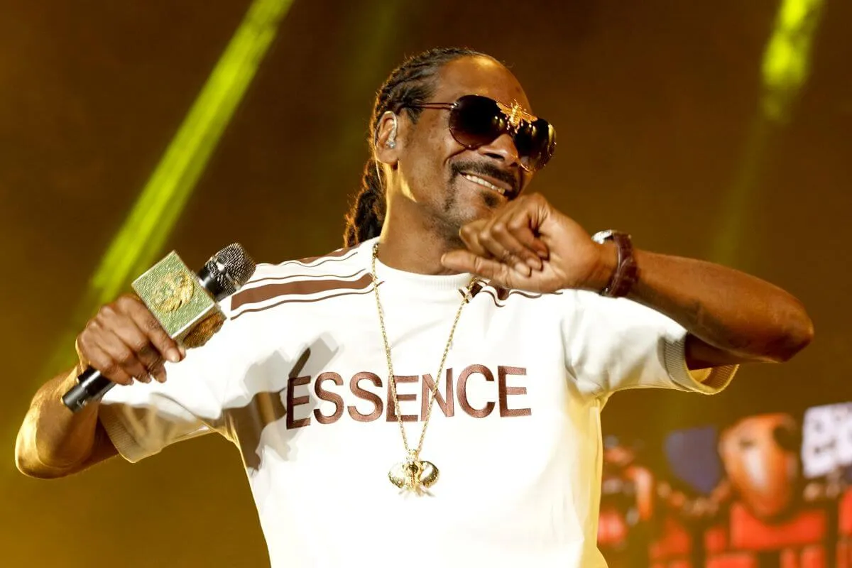 Snoop Dogg wears a white shirt and sunglasses and holds a microphone.