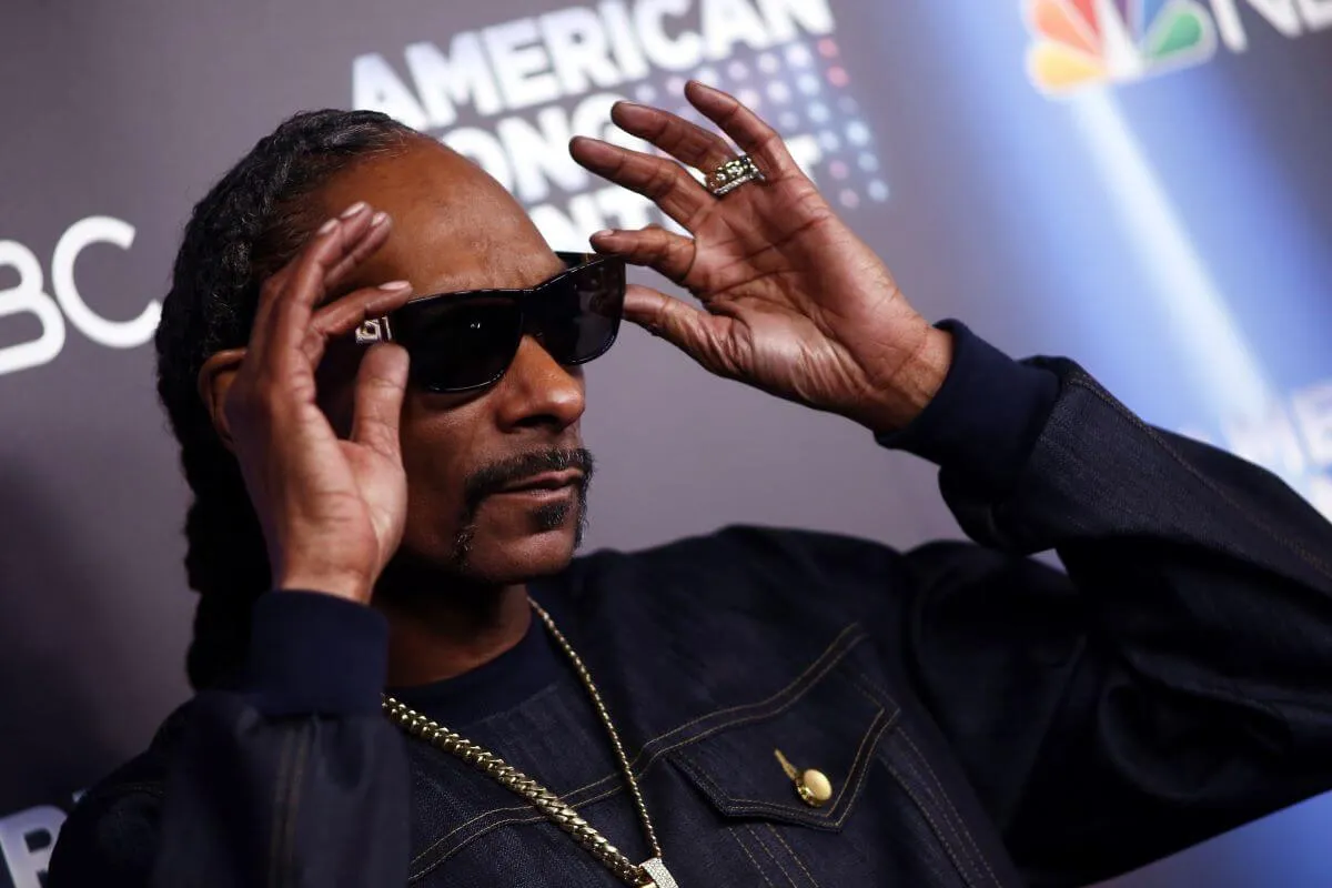 Snoop Dogg wears a black jacket and holds his hands up to his sunglasses.