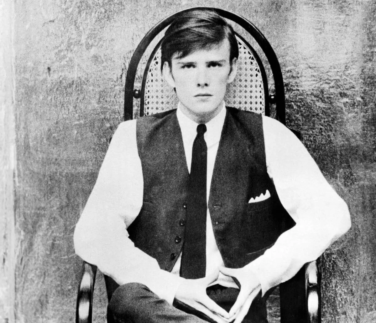 A black and white picture of Stuart Sutcliffe wearing a tie and sitting in a chair.