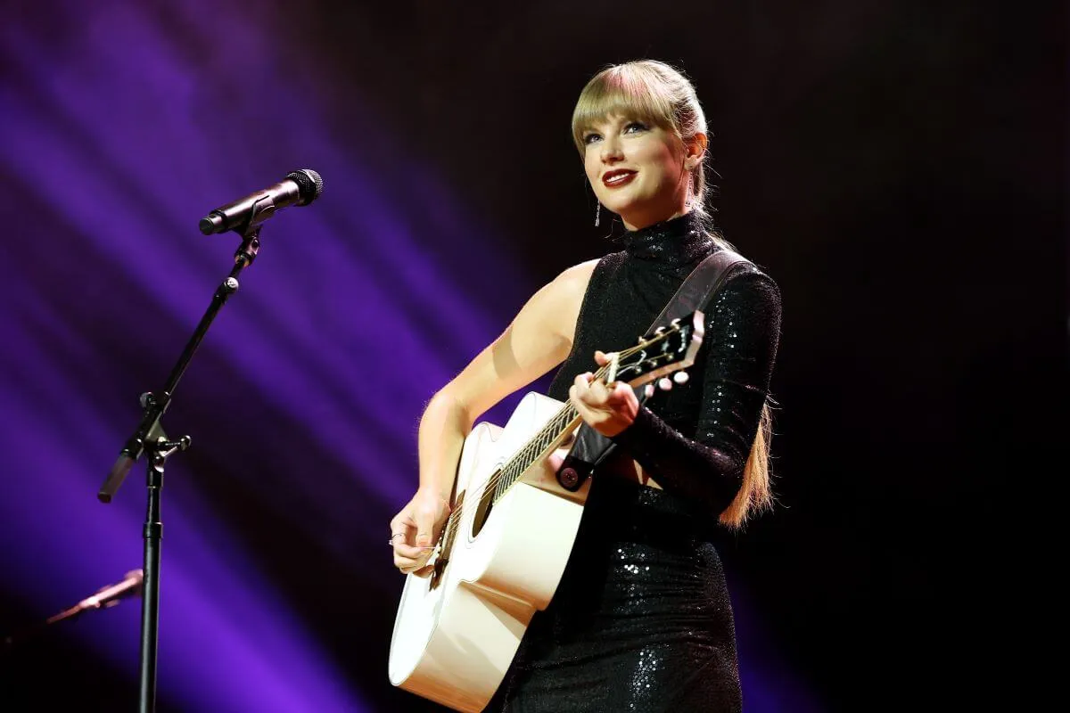 Taylor Swift wears a black to and plays a white guitar. She stands in front of a microphone.