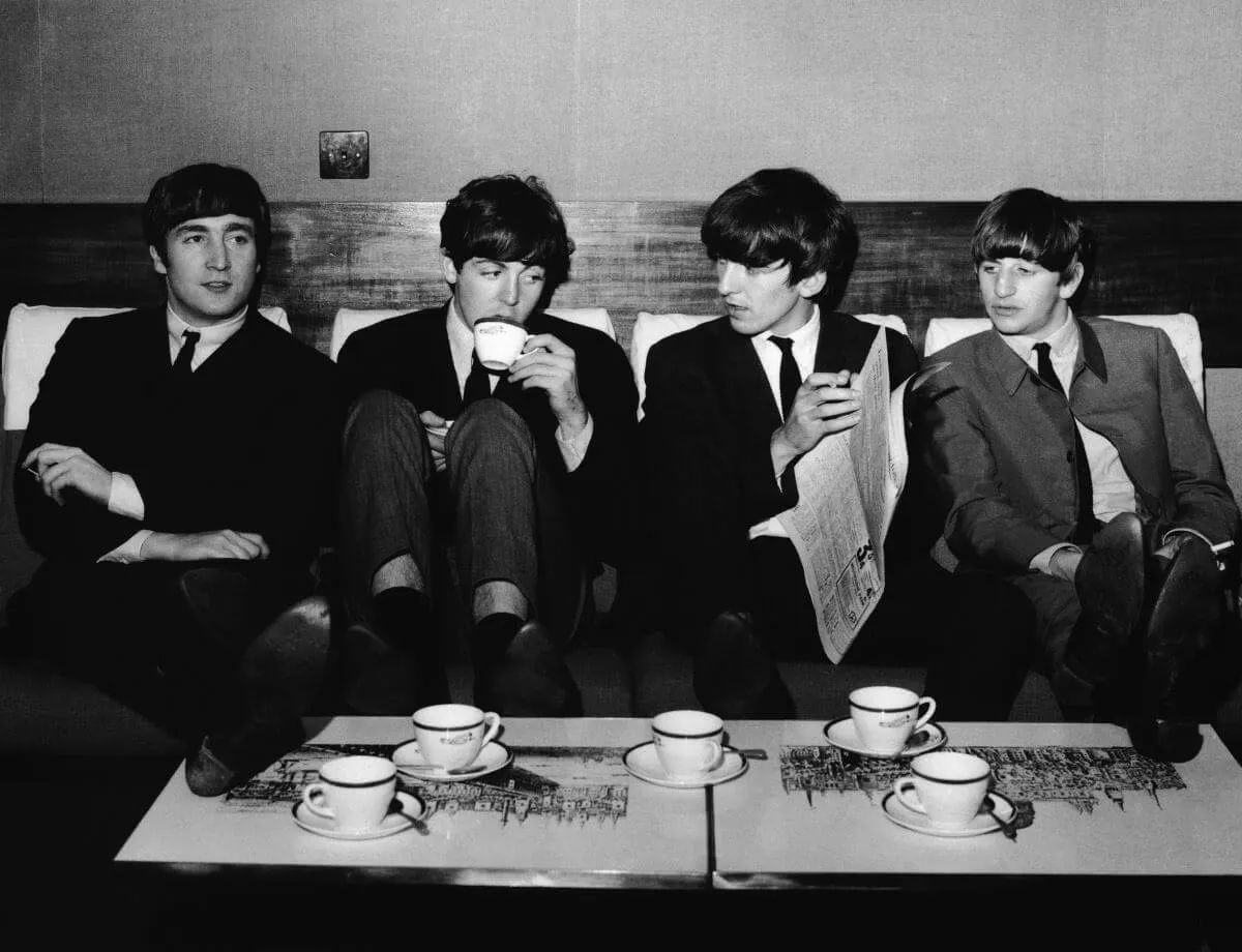 The Beatles sit on a couch together and drink tea.