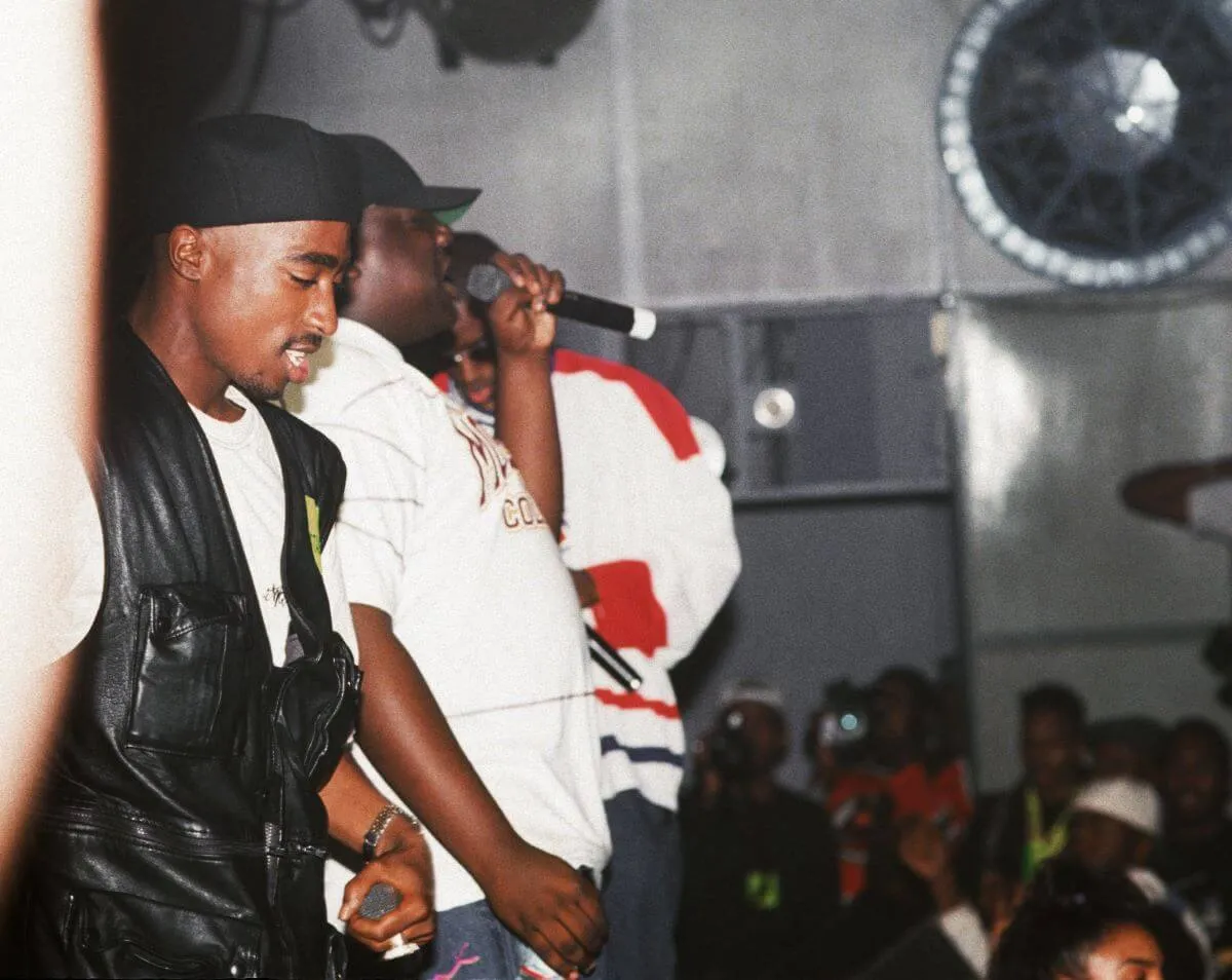 Tupac Shakur, The Notorious B.I.G., and Sean 'Diddy' Combs stand on a stage. Biggie holds a microphone.