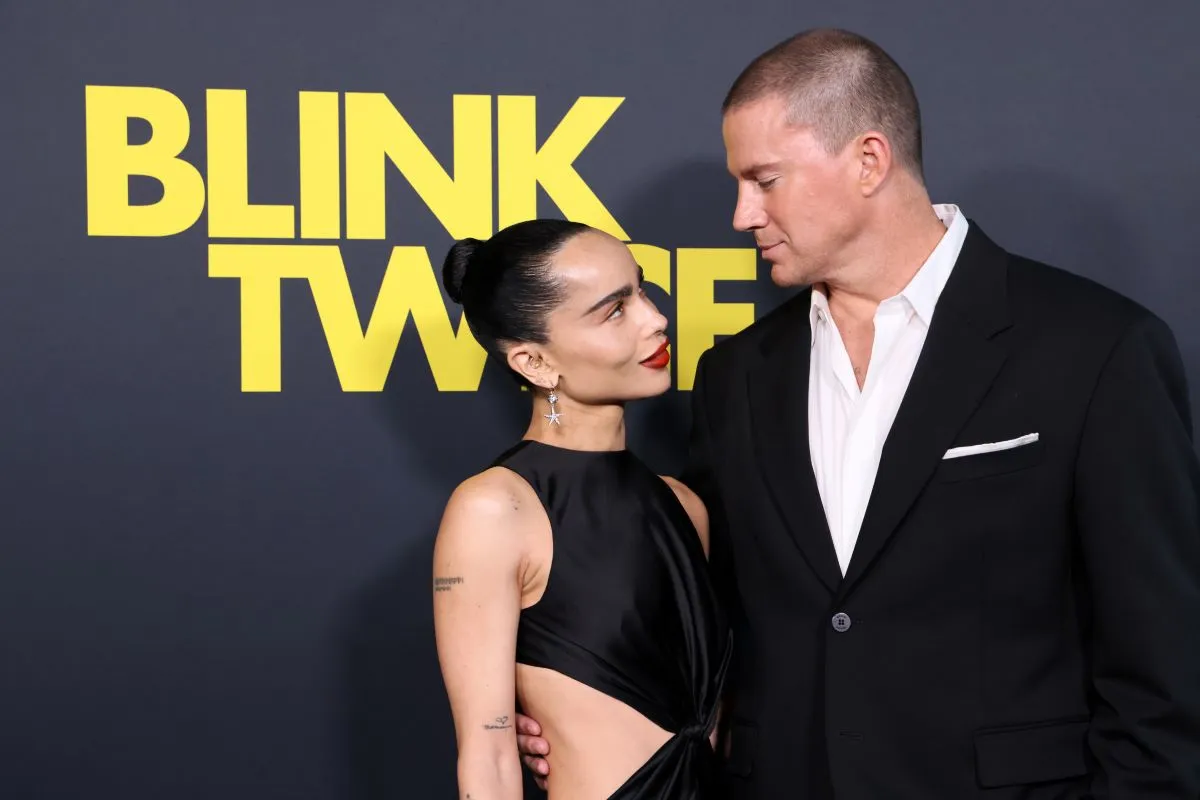 Zoe Kravitz and Channing Tatum both wear black and stand in front of a sign for 'Blink Twice.' They look at each other.