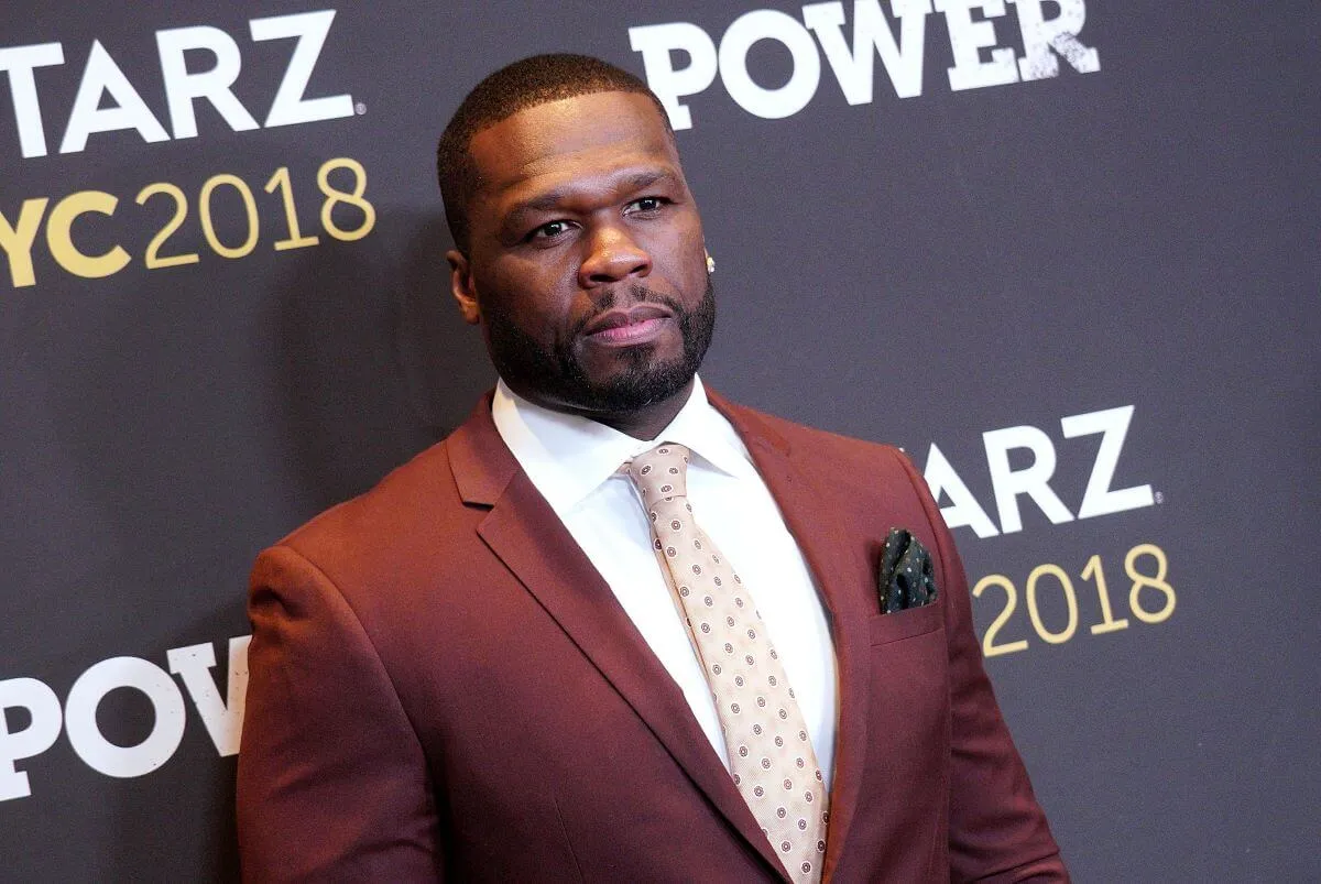 50 Cent wears a brown suit and stands in front of a black background.