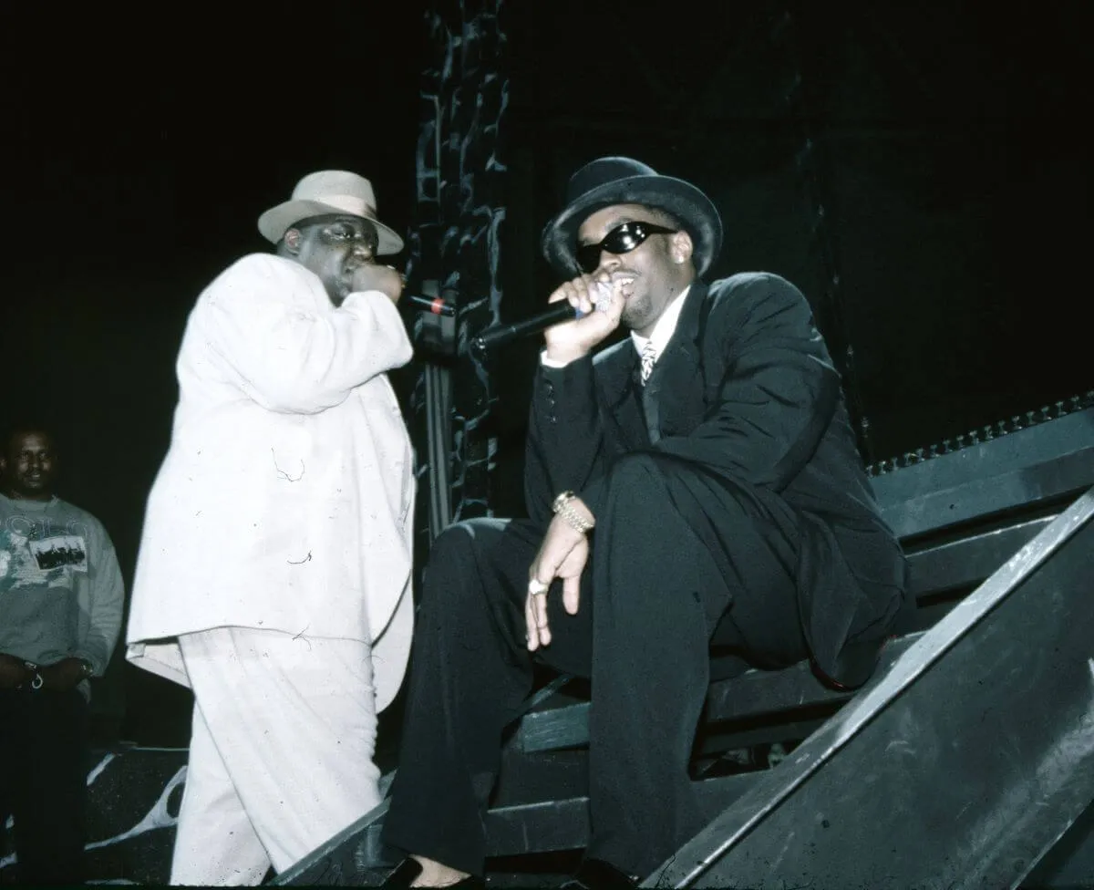 The Notorious B.I.G. wears a white suit and hat and holds a microphone. Sean 'Diddy' Combs wears a black suit and hat and sits while holding a microphone.