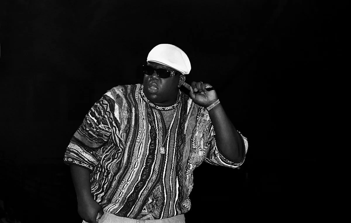 A black and white picture of the Notorious B.I.G. holding his hand to his ear. He wears a sweater, sunglasses, and a hat.