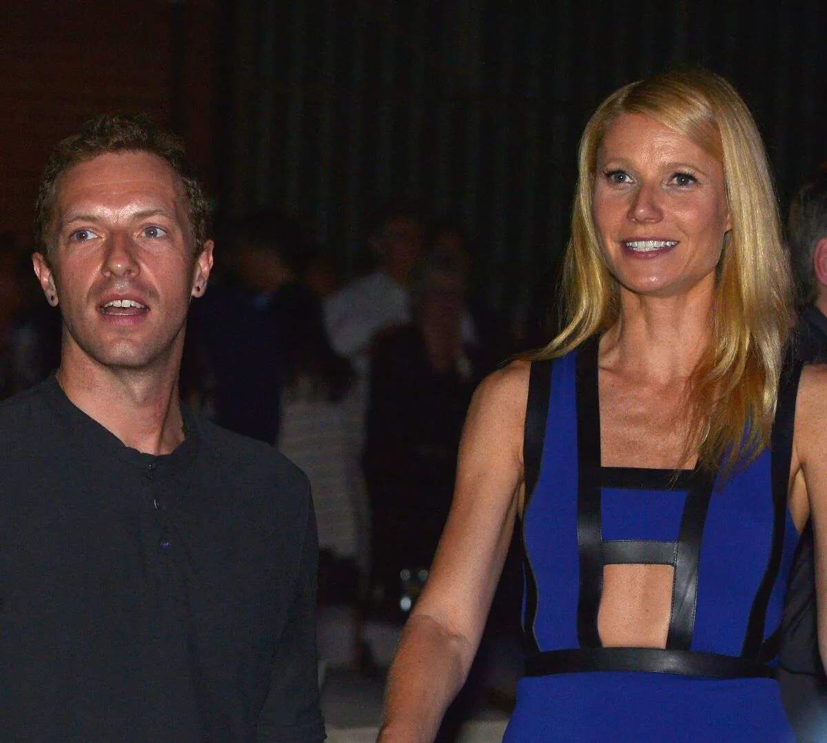 Chris Martin wears a black shirt and stands with Gwyneth Paltrow, who wears a blue and black dress.