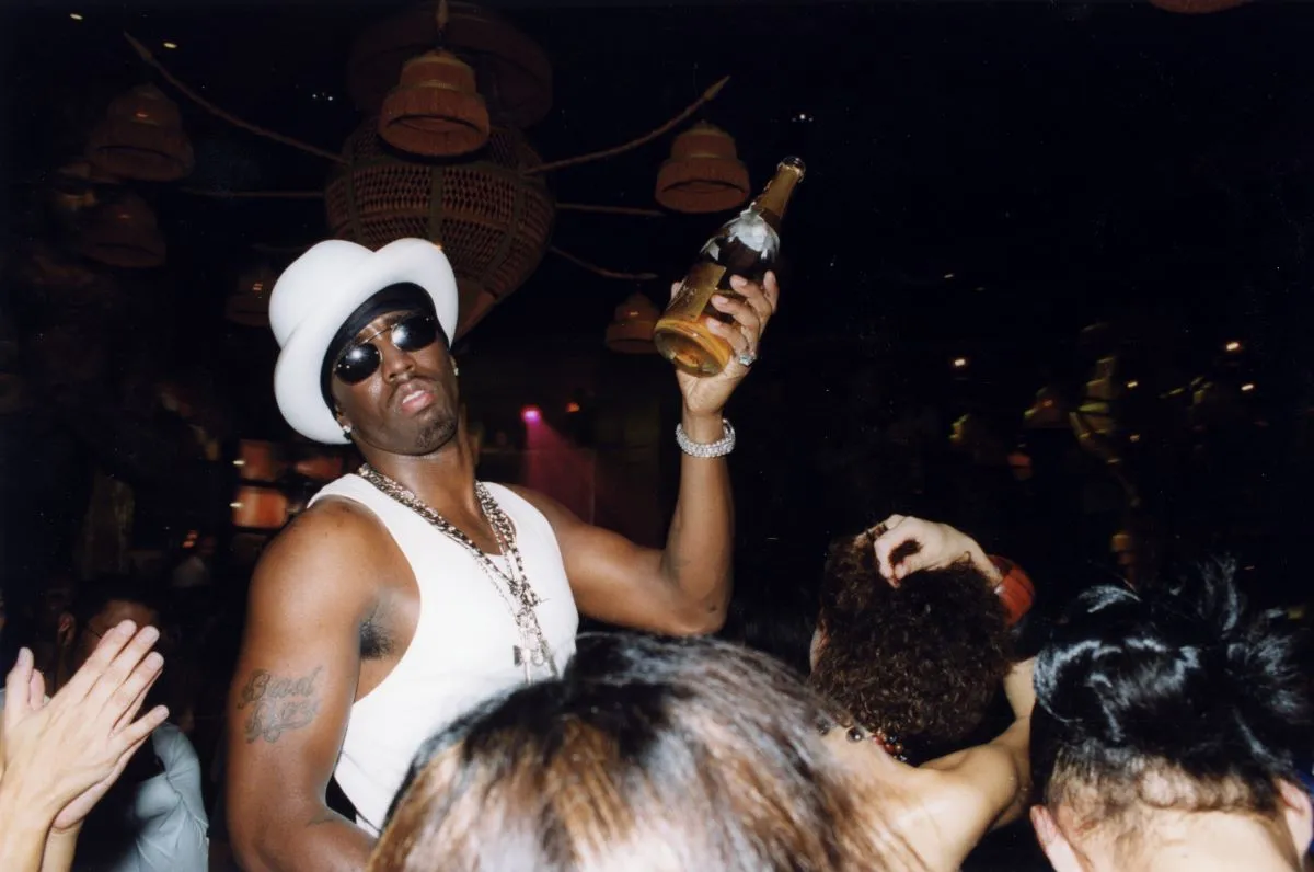 Sean 'Diddy' Combs wears a white hat and sunglasses. He holds a bottle of champagne and stands in a crowd.