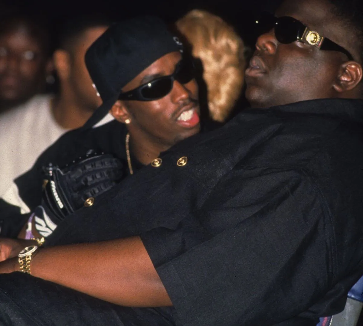 Sean 'Diddy' Combs wears a hat and sunglasses and talks to the Notorious B.I.G., who wears sunglasses. They both wear black.