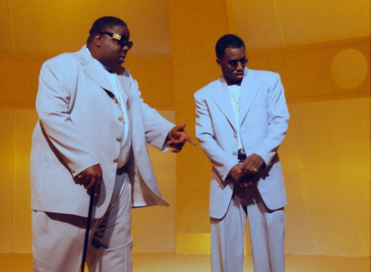 The Notorious B.I.G. and Sean 'Diddy' Combs wear white suits and stand against a yellow background.