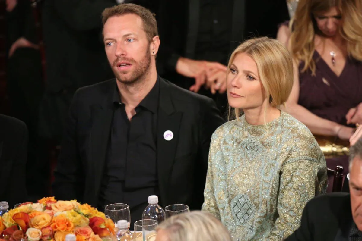 Chris Martin and Gwyneth Paltrow sit together at a table. He wears a black suit and she wears a green dress.