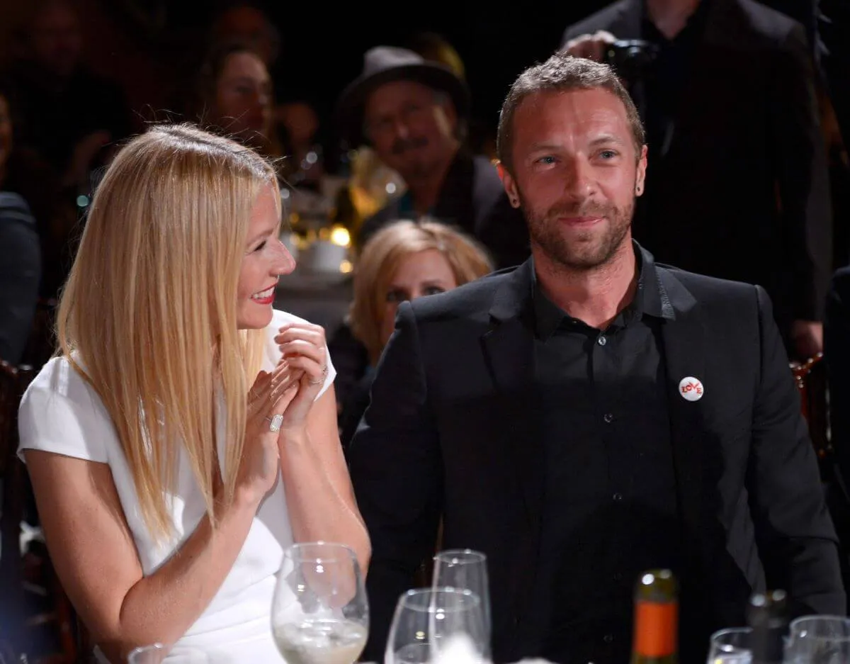 Gwyneth Paltrow wears a white shirt and smiles while she looks at Chris Martin. He wears black. They sit at a table.