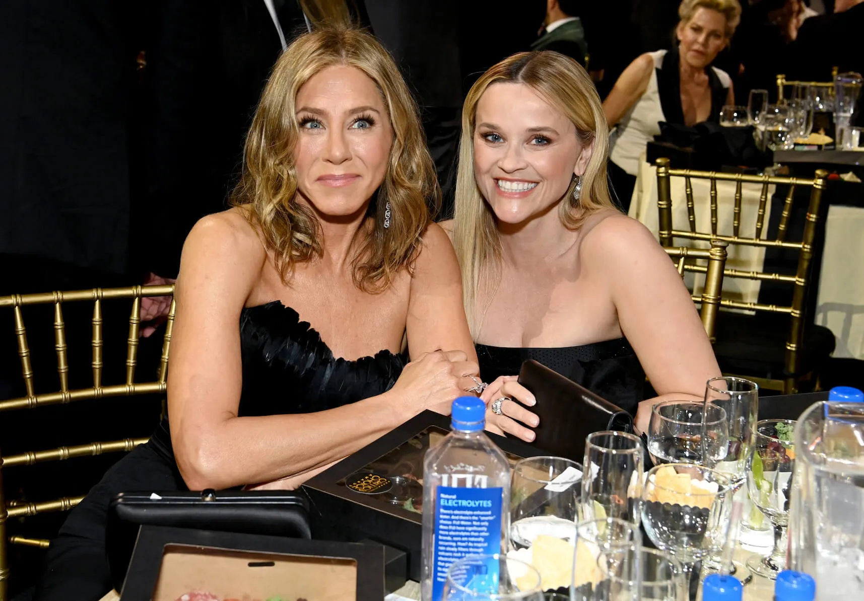 Jennifer Aniston and Reese Witherspoon sitting next to each other arm in arm and wearing black sleeveless dresses at the Critics Choice Awards in 2024