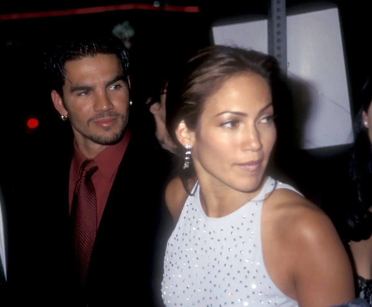 Jennifer Lopez and her first husband, Ojani Noa, stand together and look to the side. He wears a red shirt and she wears a silver dress.