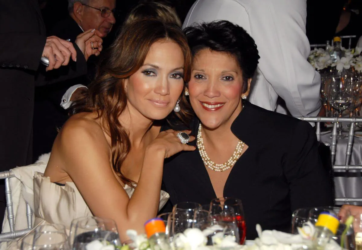 Jennifer Lopez wears a white dress and leans into her mom, who wears black. They both sit at a table.