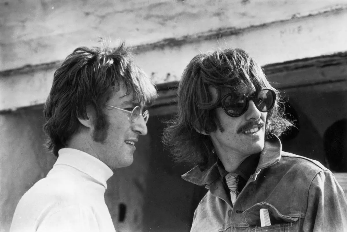 A black and white picture of John Lennon and George Harrison standing outside. Lennon wears a turtleneck and glasses and Harrison wears a denim jacket and sunglasses.