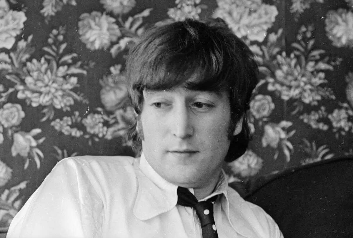 A black and white picture of John Lennon wearing a shirt and tie. He sits in front of a wall with floral wallpaper.