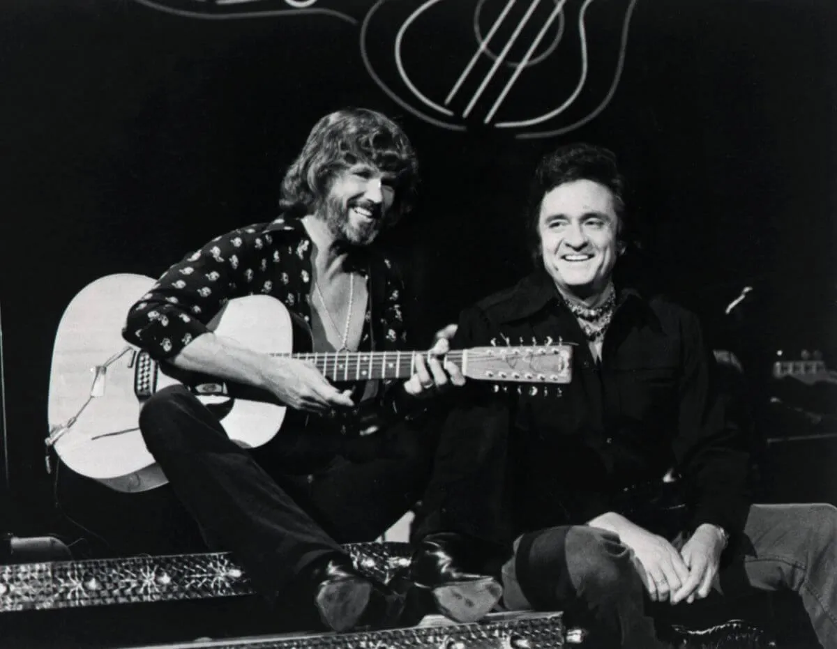 A black and white picture of Kris Kristofferson and Johnny Cash sitting together. Kristofferson holds a guitar.