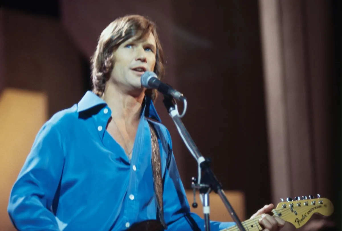 Kris Kristofferson wears a blue shirt and sings into a microphone.