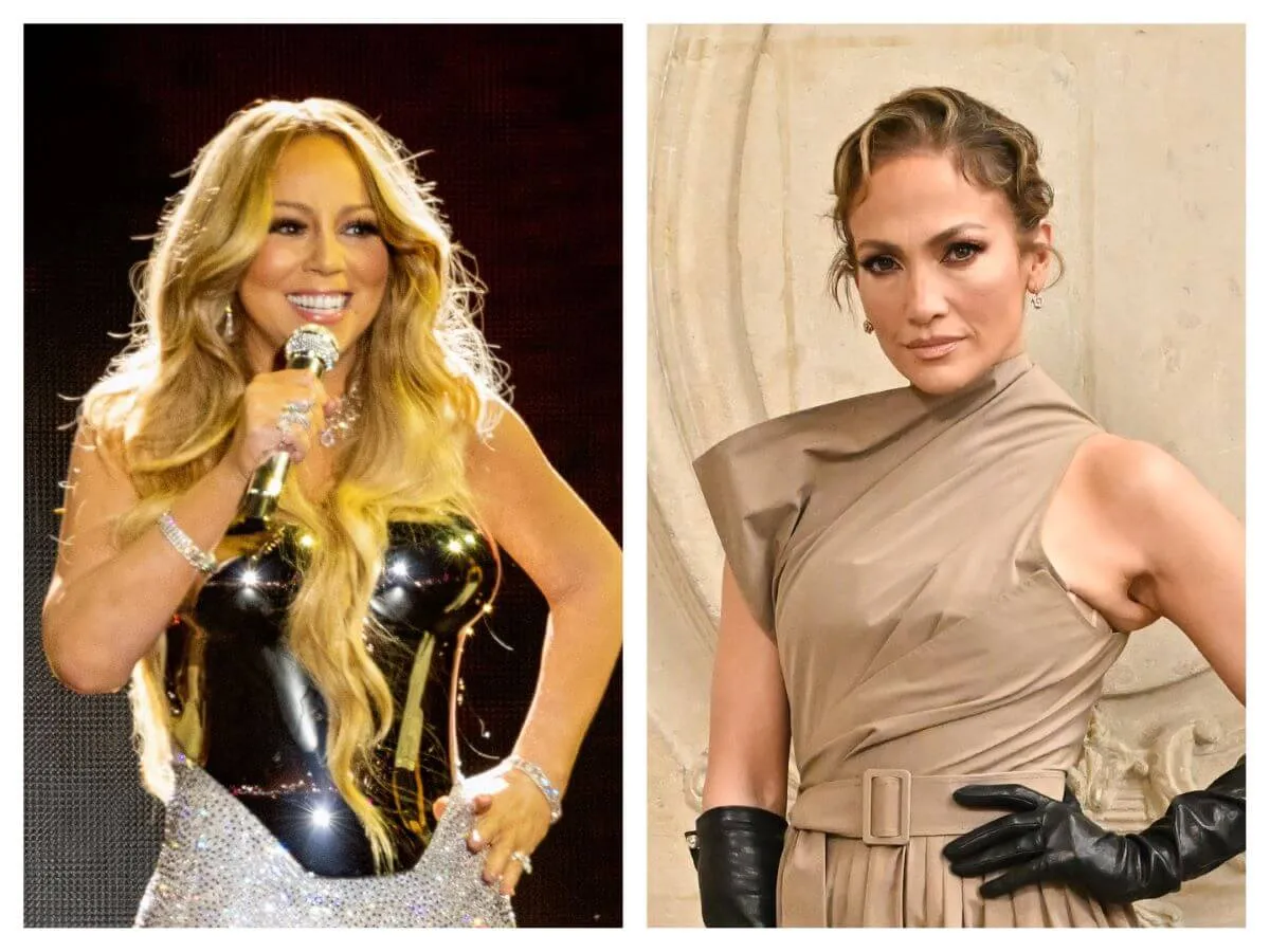 Mariah Carey stands on stage with one hand on her hip. She holds a microphone to her face with the other hand. Jennifer Lopez wears black leather gloves and stands with her hand on her hip.