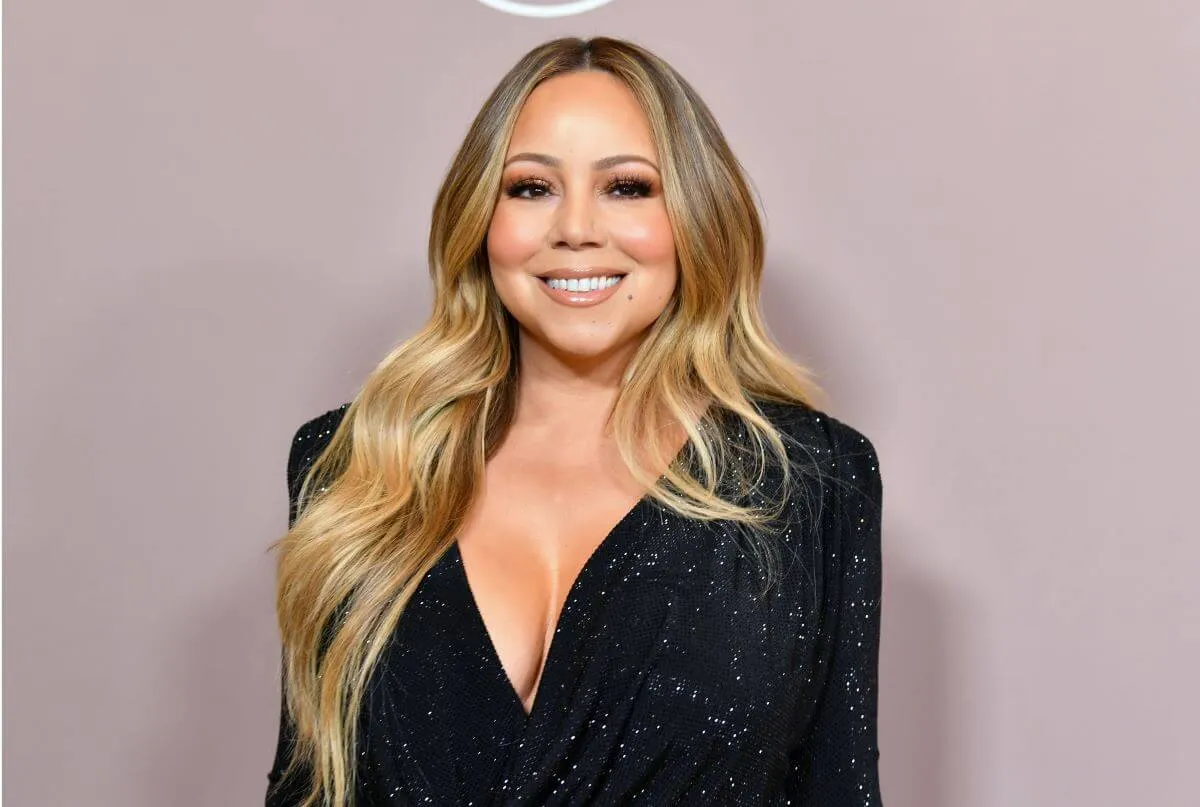 Mariah Carey wears a black dress and stands against a light pink wall.