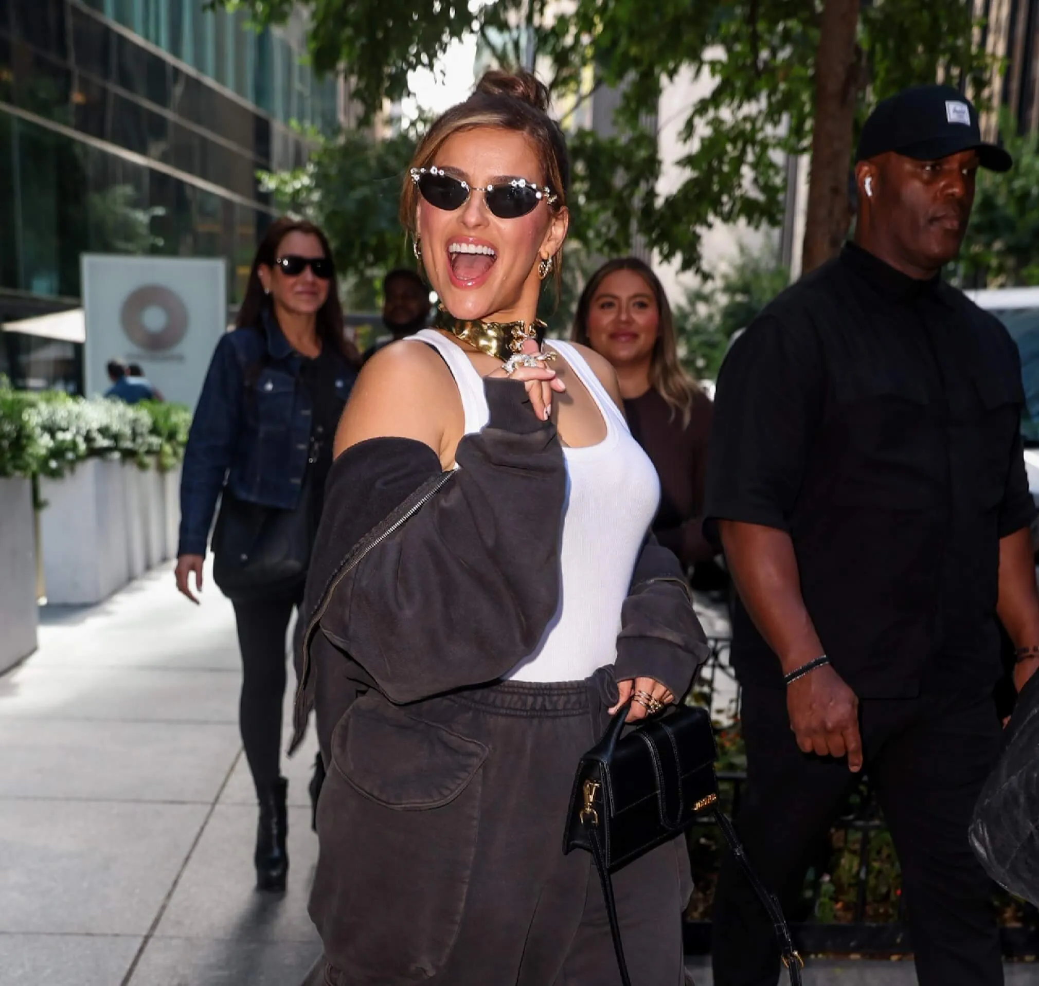 Nelly Furtado wears a white tank top and sunglasses. She waves at the camera.