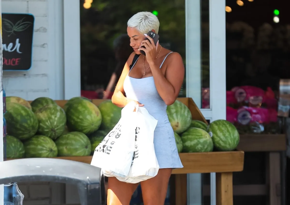 Wearing a blue tennis dress, Nicole Mitchell exits a grocery store carrying two grocery bags and talking on her cell phone