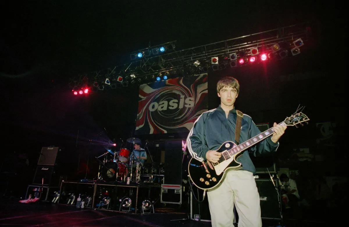 Noel Gallagher stands onstage at an Oasis concert and holds a guitar.