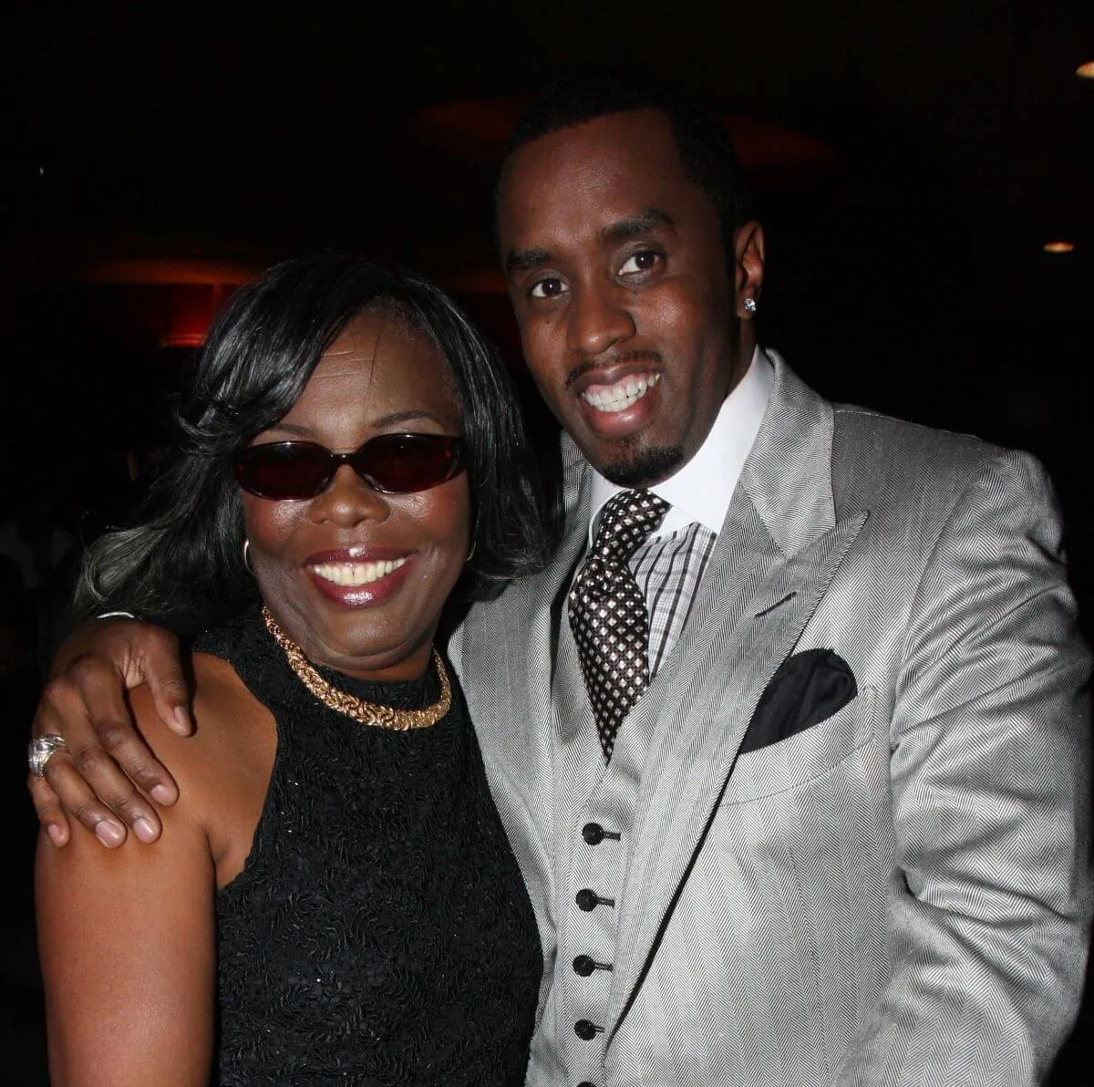 Diddy stands with his arm around the Notorious B.I.G.'s mother, Voletta Wallace. She wears a black dress and sunglasses and he wears a silver suit.