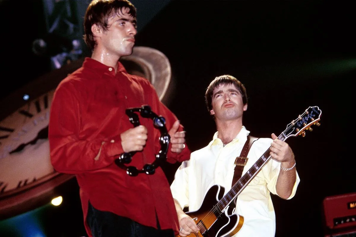 Liam and Noel Gallagher of Oasis stand on a stage. Liam wears a red shirt and plays a tambourine. Noel wears a white shirt and plays guitar.