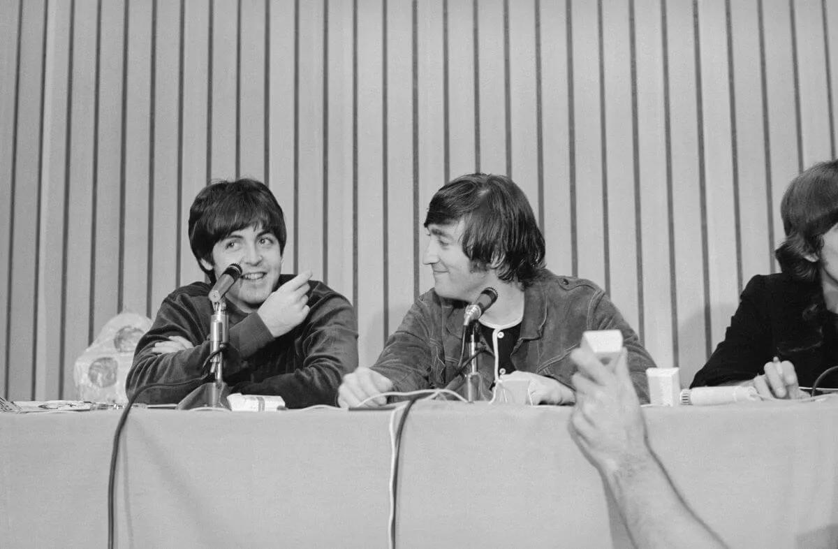 A black and white picture of Paul McCartney and John Lennon sitting behind a table together. There are microphones in front of them.