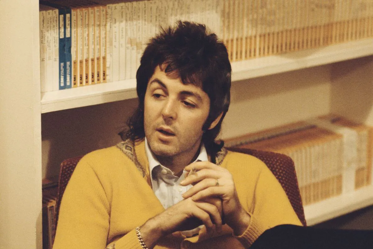 British musician Paul McCartney wears a yellow sweater and sits in front of some shelves.