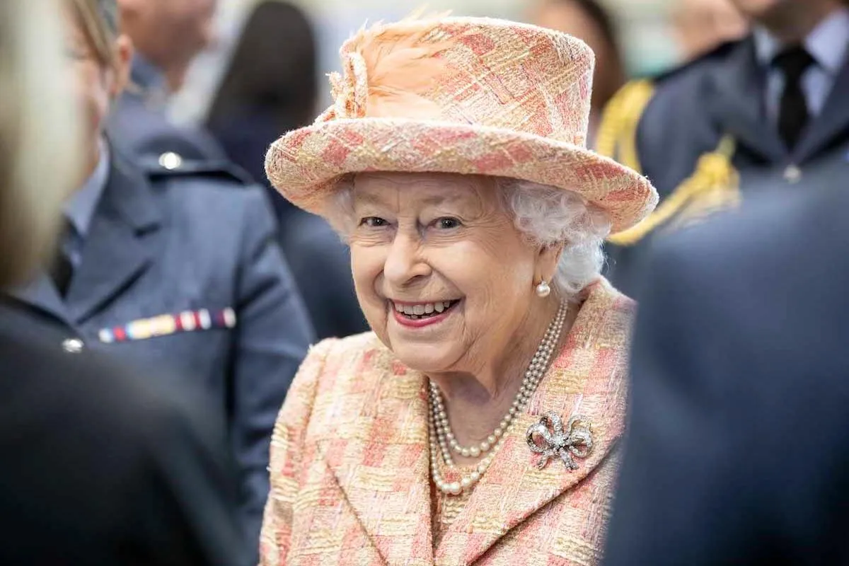 Queen Elizabeth II smiles and looks on wearing a peach-colored suit and hat.