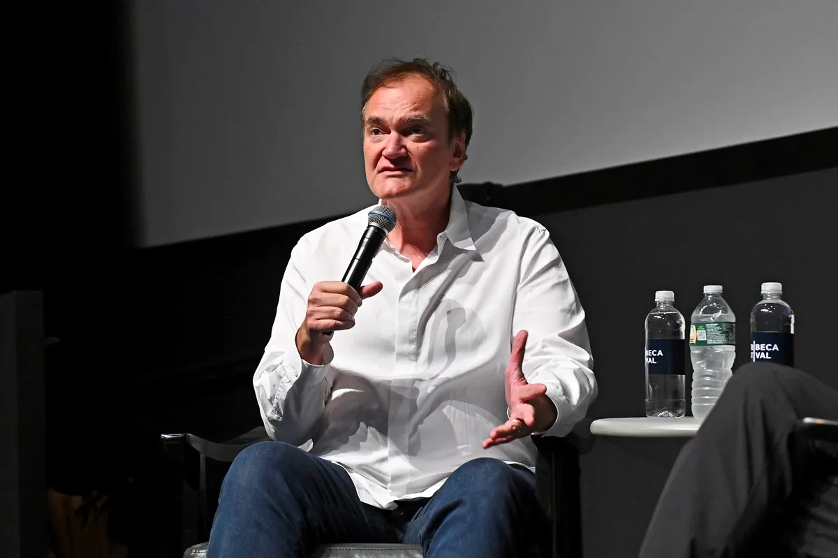 Quentin Tarantino speaks onstage at the "Jackie Brown" screening during the 2024 Tribeca Festival while wearing a white button-up shirt and blue jeans.