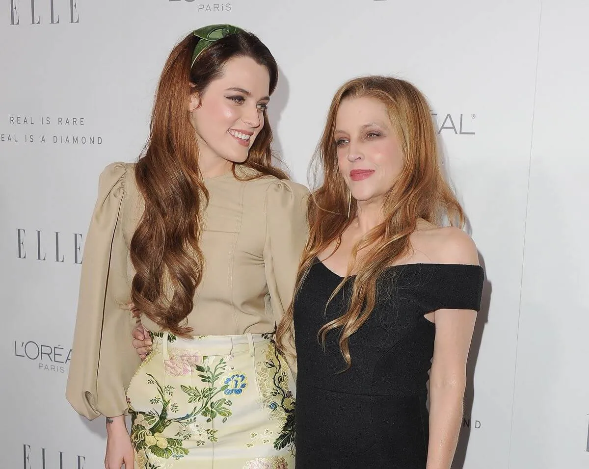 Riley Keough and Lisa Marie Presley stand together. Keough wears a skirt and sweater and Presley wears a black dress.