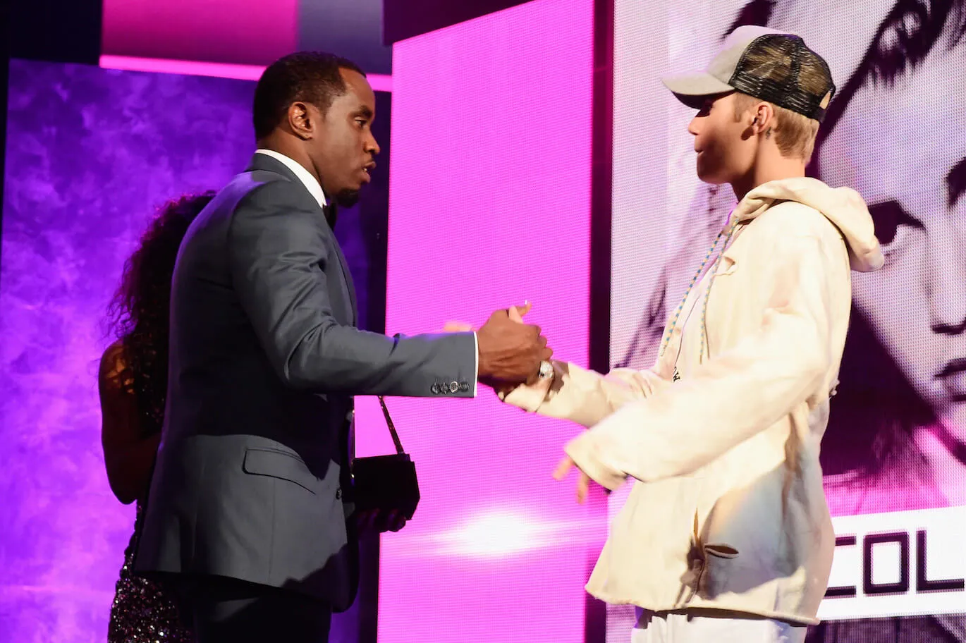 Sean 'Diddy' Combs and Justin Bieber shaking hands at an awards ceremony. They're against a pink and purple background.