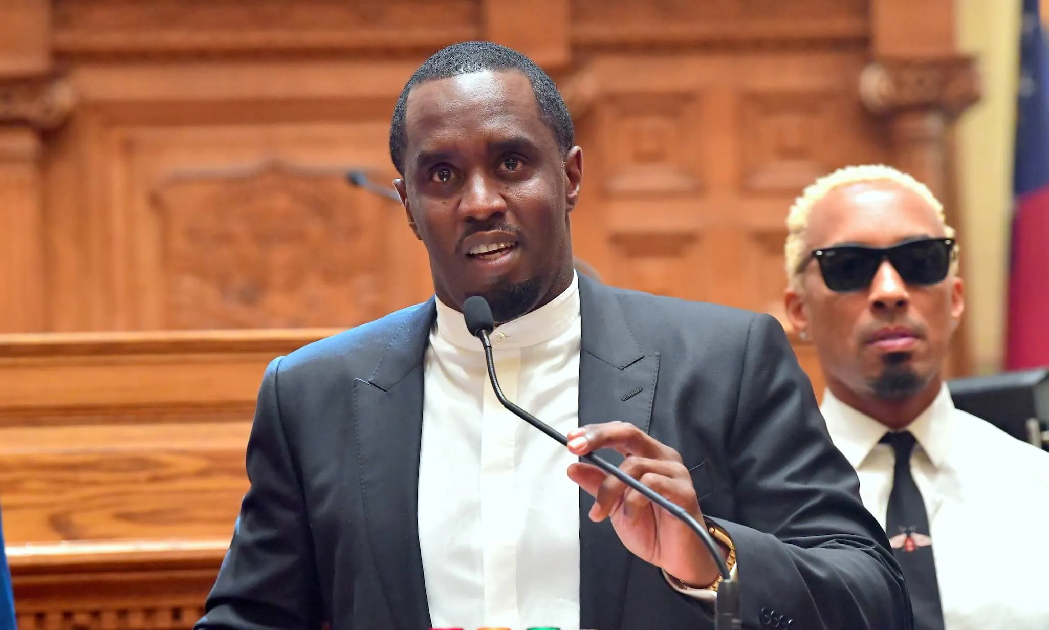 Sean 'P. Diddy' Combs in a suit over a white shirt while speaking into a microphone attached to a stand