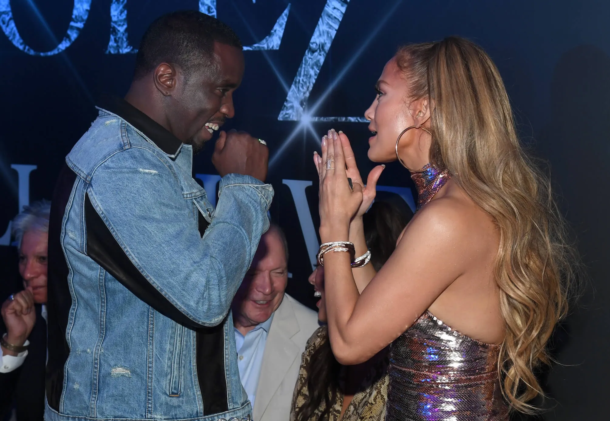 Sean 'P. Diddy' Combs and Jennifer Lopez facing each other looking excited. Combs is smiling with his fists under his chin. Lopez is holding her hands together like she's praying in front of her face.