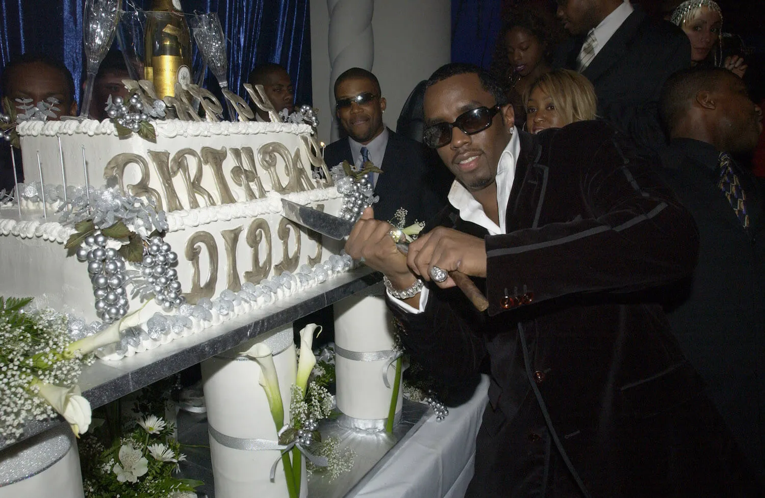 Sean 'P. Diddy' Combs holding a cake cutter up to his giant birthday cake that says "Birthday Diddy." He's wearing a suit and sunglasses.