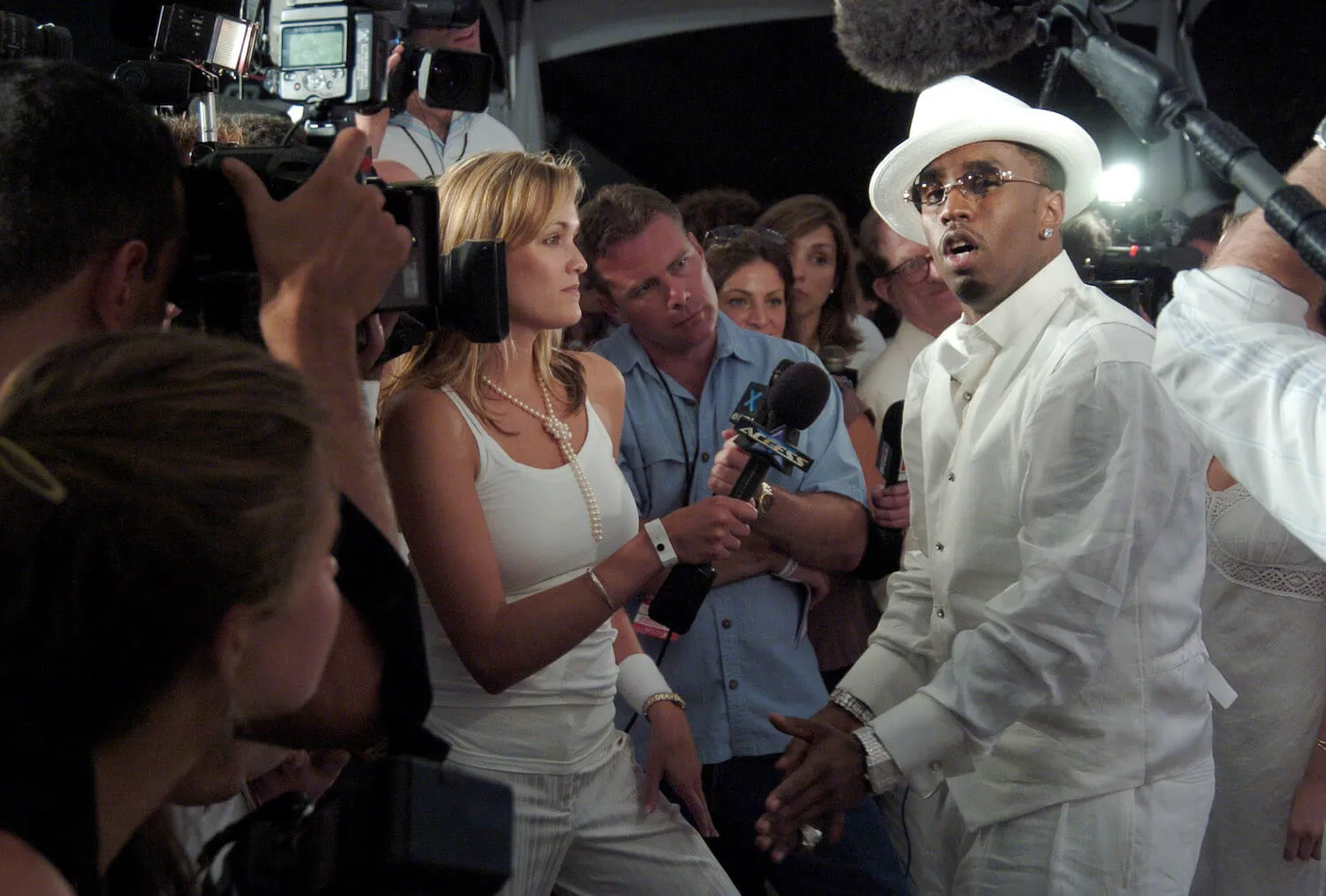 Sean 'P. Diddy' Combs  speaking into a microphone at one of his white parties in the Hamptons. He's wearing a white suit and hat. He's surrounded by reporters. 