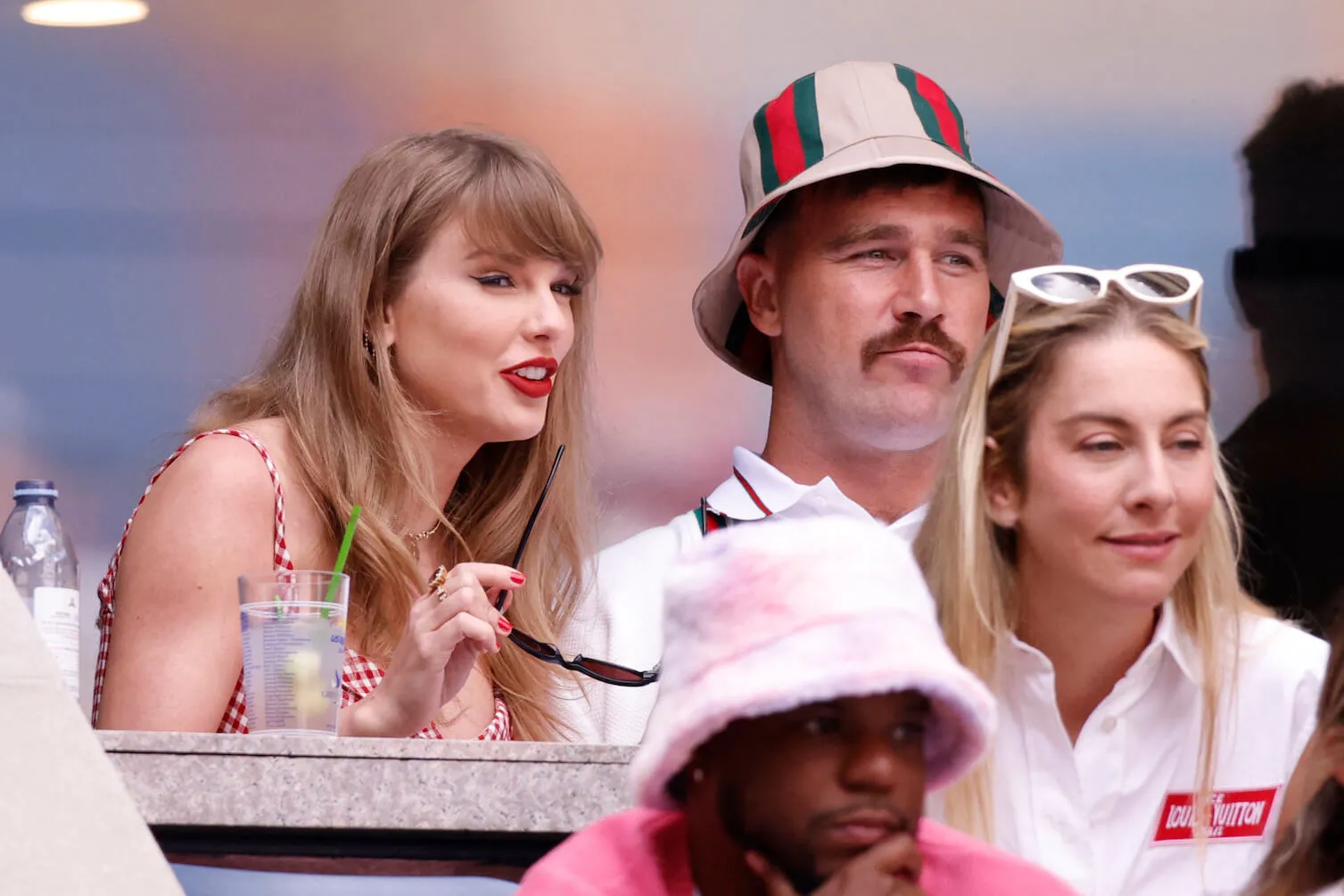 Taylor Swift and Travis Kelce sitting next to each other at the US Open. Swift is holding her sunglasses and wearing a plaid dress. Kelce is wearing a striped bucket hat.